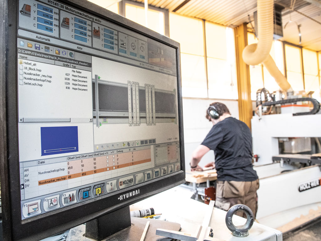 Im Vordergrund steht ein Computerbildschirm mit einer digitalen Darstellung eines Holzbretts und dessen Maßen. Im Hintergrund schneidet ein Arbeiter ein Holzbrett an einer Maschine passend zu.