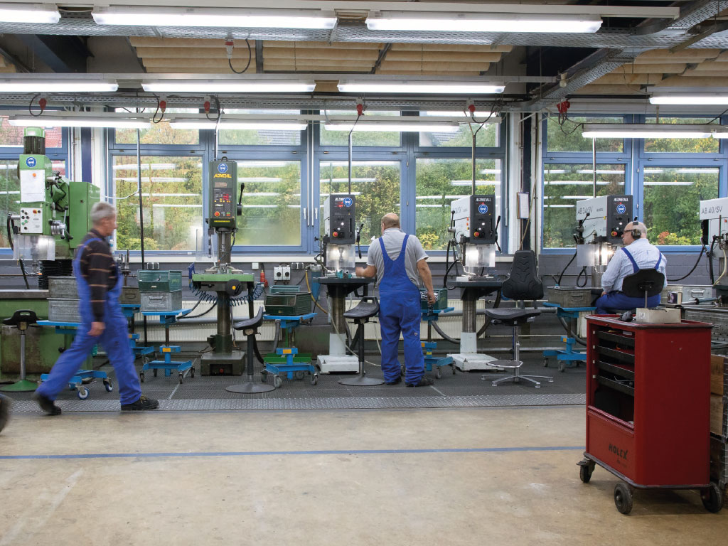 In einer großen Halle stehen mehrere Maschinen nebeneinander. Davor stehen oder sitzen Menschen, die an den Maschinen arbeiten.